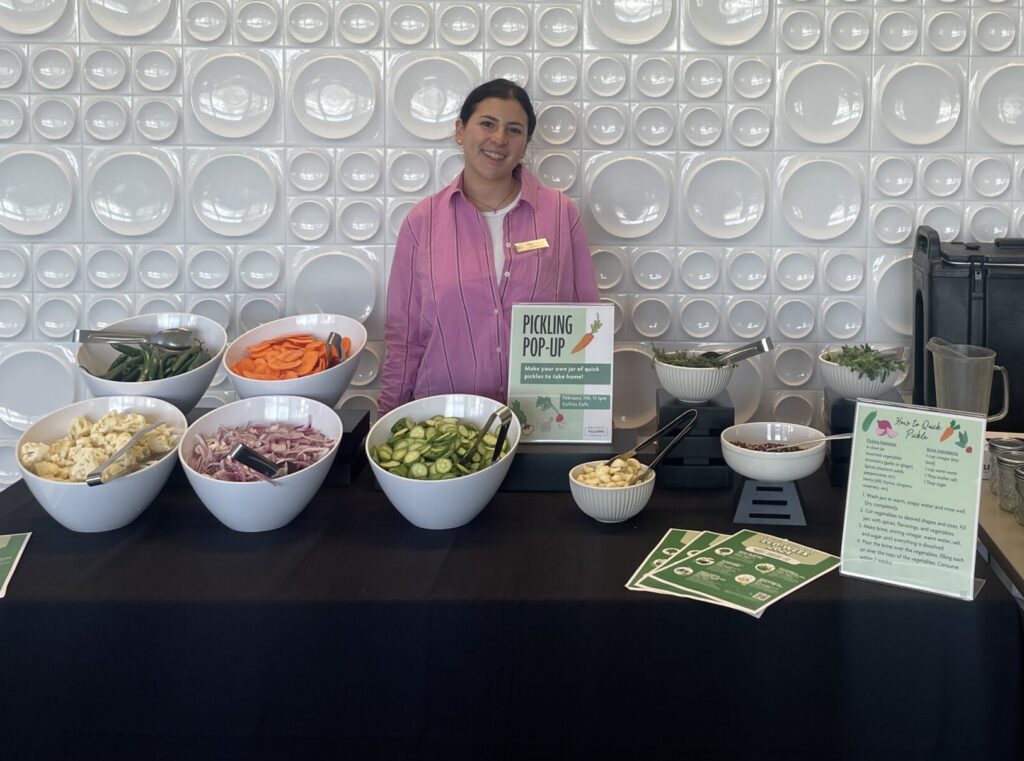 A young woman with brown hair stands behind a table displaying ingredients for a pickling demonstration.
