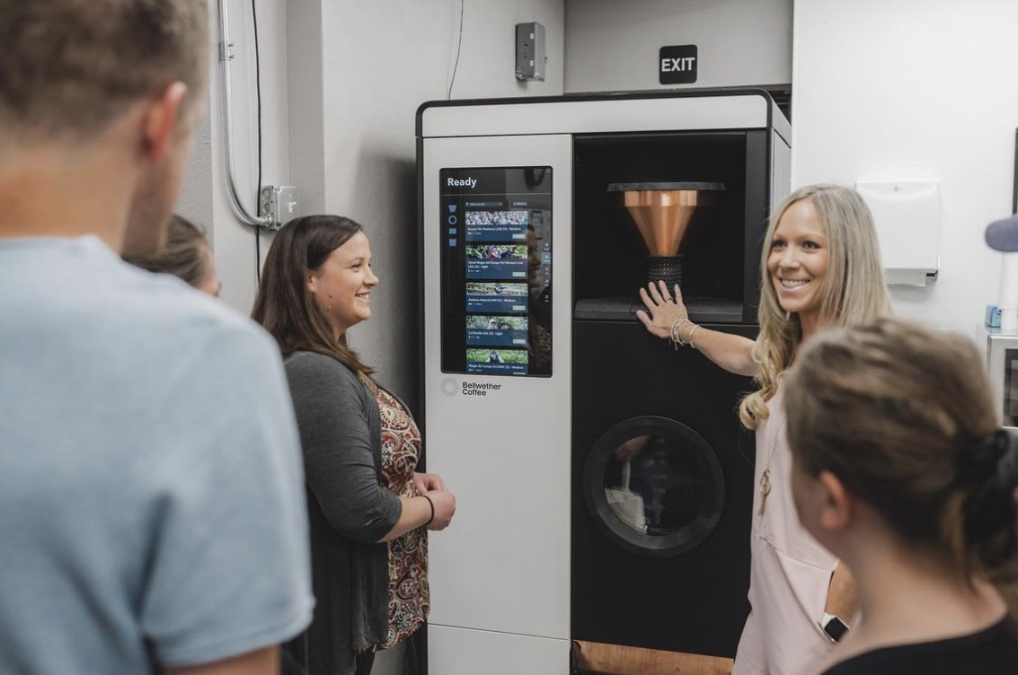 A woman demonstrates how to use a piece of coffee roasting equipment to a group of people. 