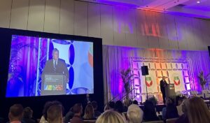 A man speaks at a podium with a large screen next to him projecting his image.