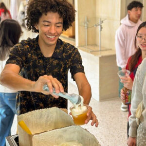 A student pours himeself apple cider from a large container with a line of people behind him. 