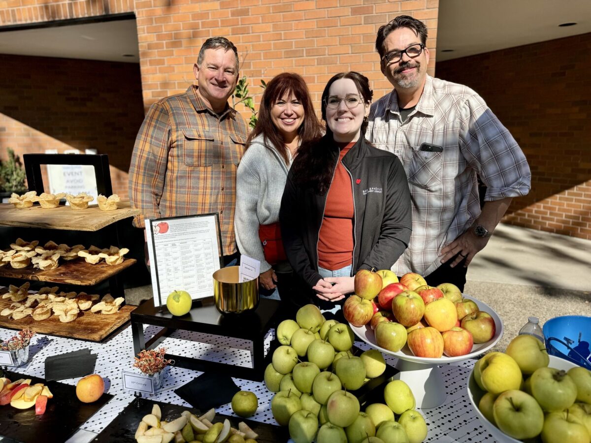 Four adults stand smiling behind a display table full of all different kinds of apples.