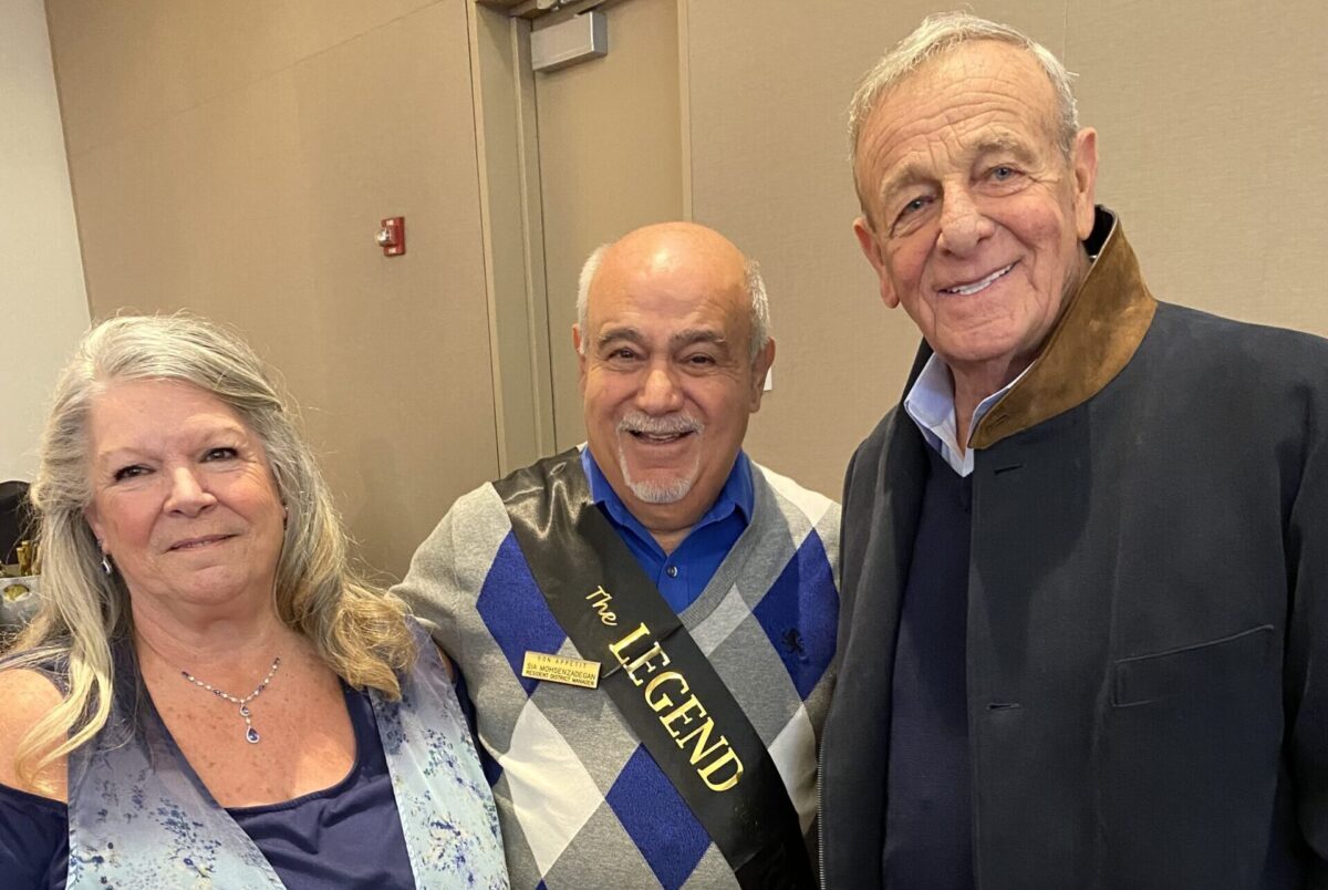 A woman and two men stand together for a portrait. The man in the center is wearing an argyle sweater and a banner that says, "the legend."