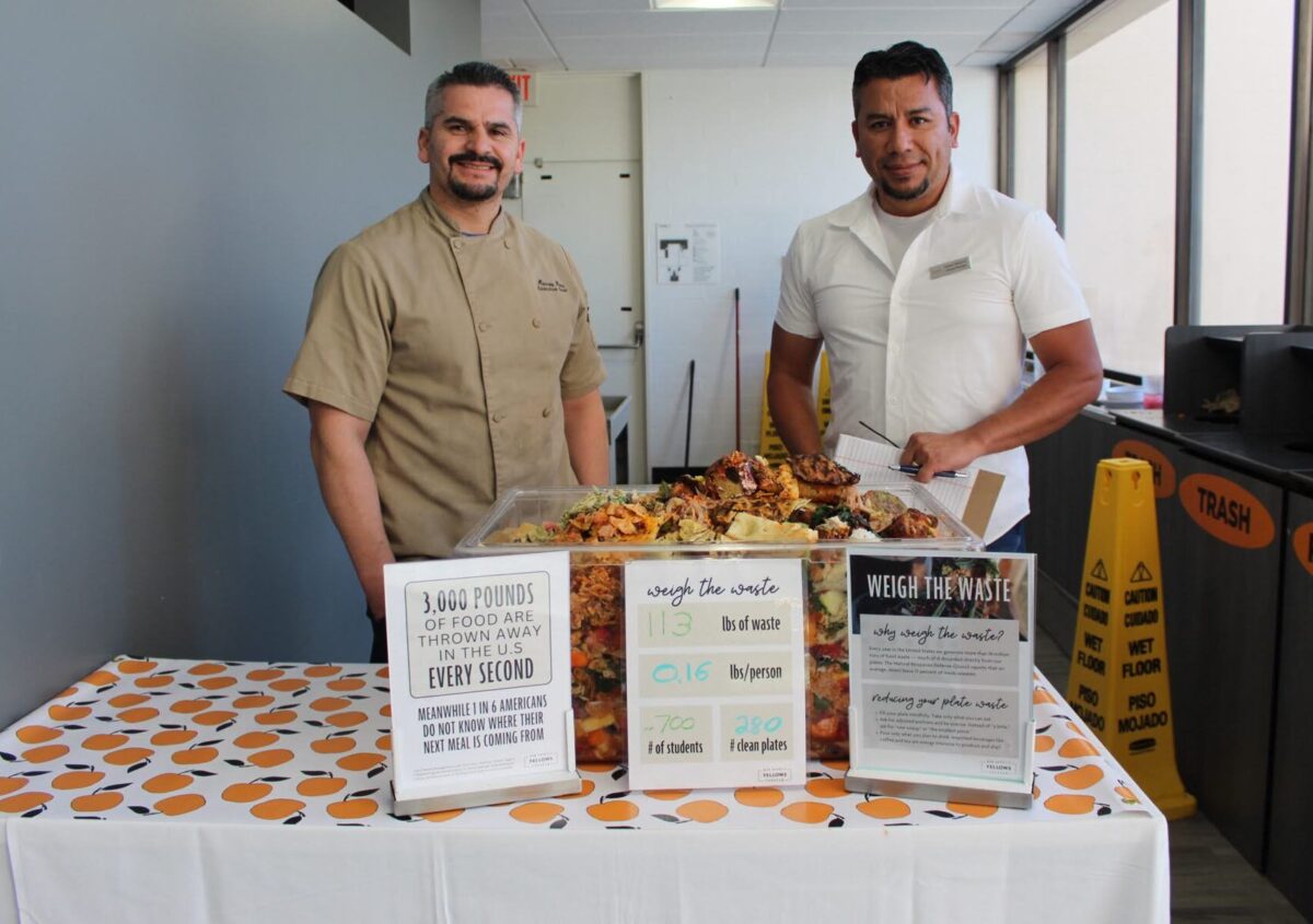 Two men in culinary uniforms stand behind a table with a bin full of food waste and three signs talking about food waste.