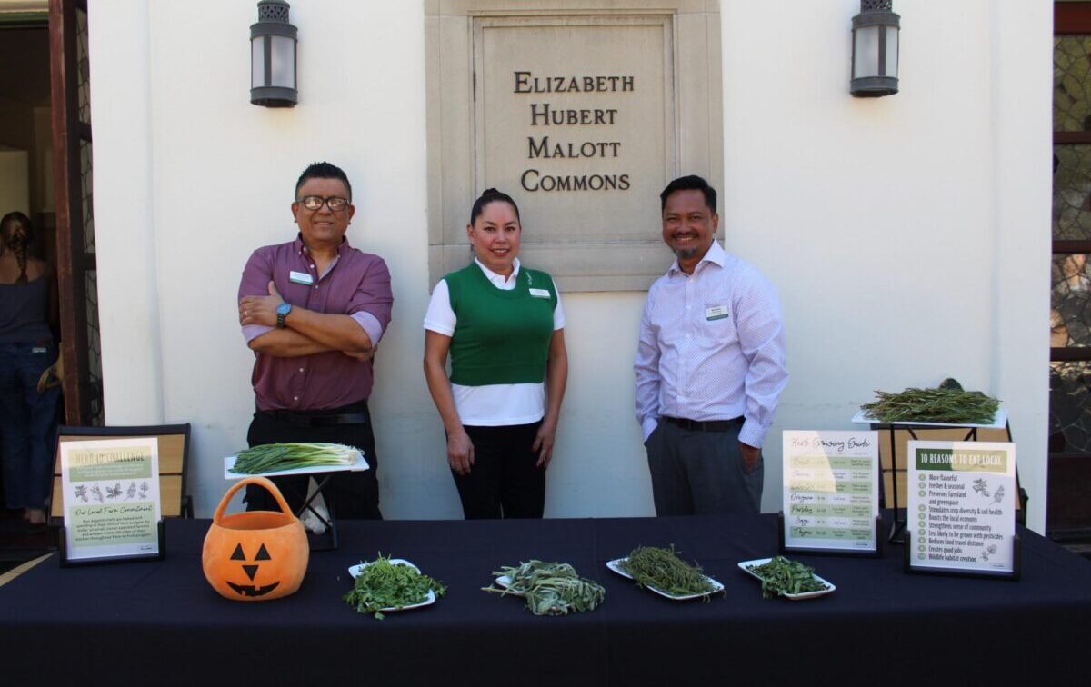 A woman in a green vest and white shirt stand between two men who at a table with piles of herbs and educational signage about herb use.
