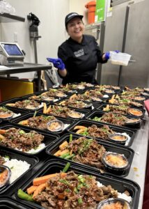 A woman chef stands in front of a table filled with rows of boxes of takeout food. 