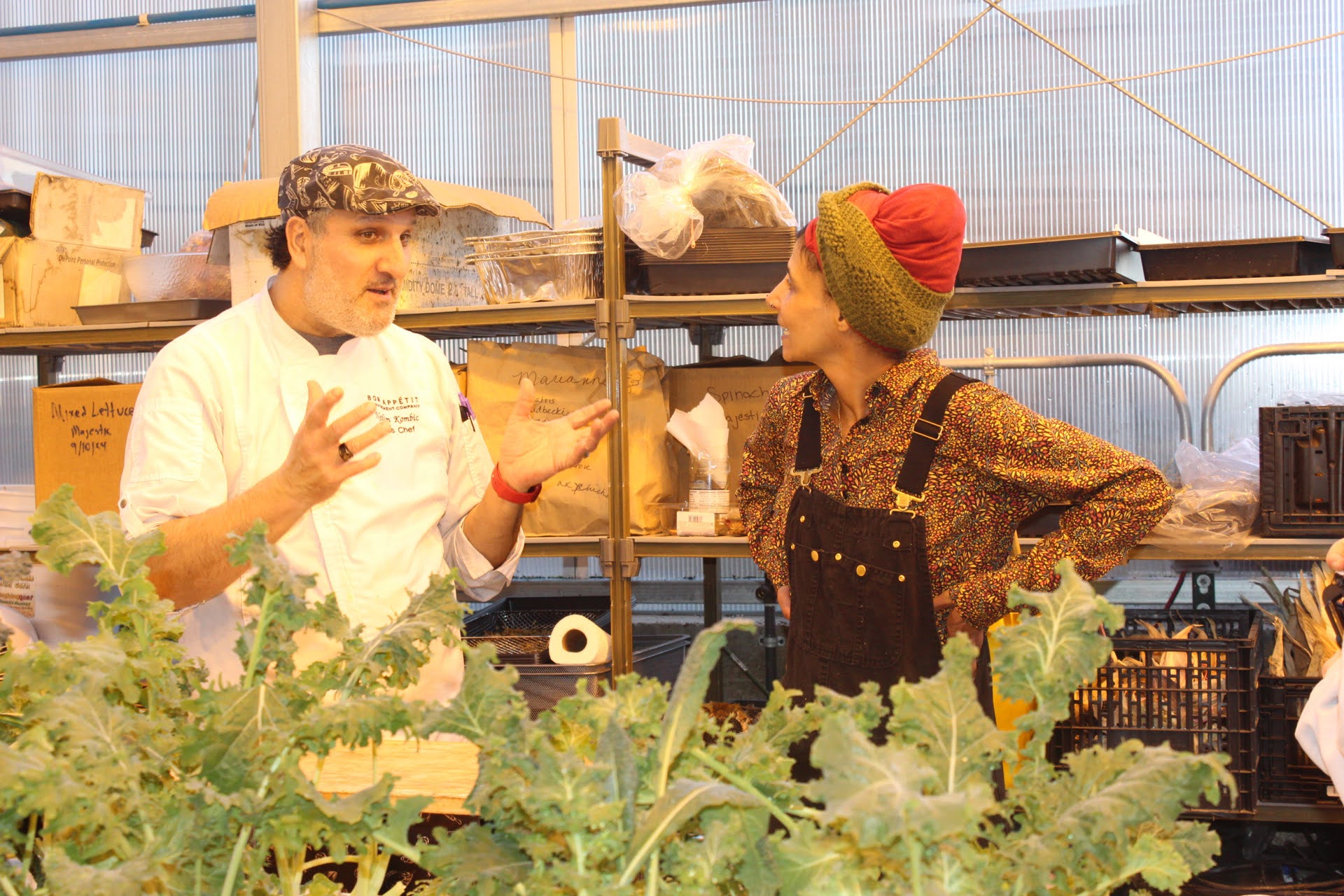 A man in a chef's coat and dark hat speaks with a woman in a kit cap and black overalls. They are in a greenhouse. 