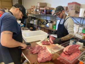 Two men trim beef in a commisary-looking kitchen. 