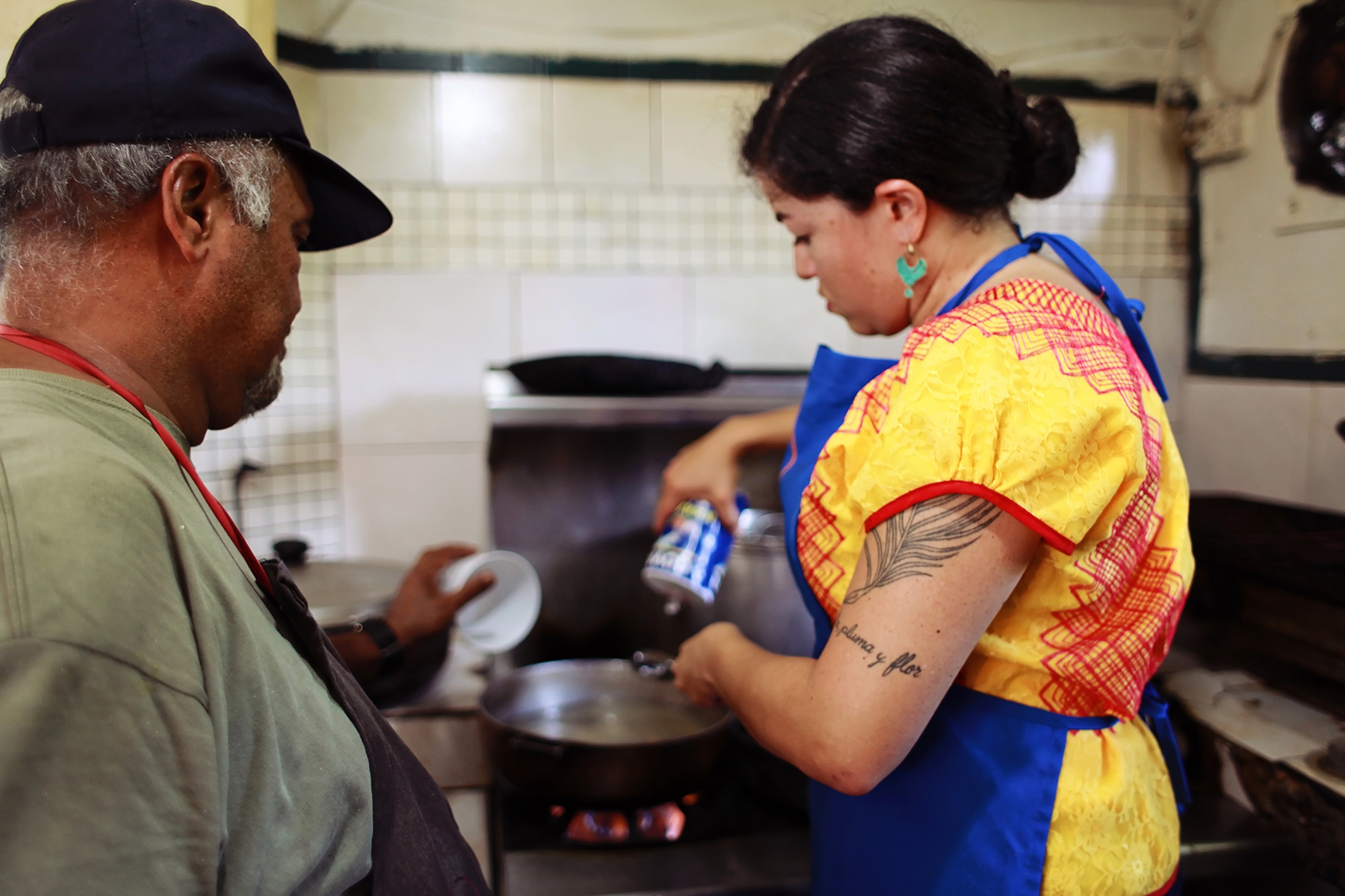 A woman in a bright yellow top and a blue apron stands at the stove, measuring an ingredient, while a man in a ball cap looks on. 